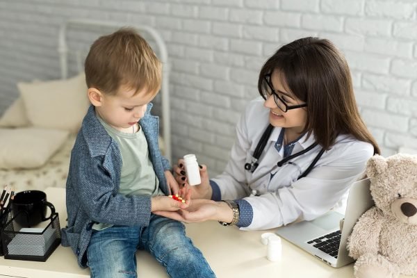 Beautiful smiling female doctor hold in arms pill bottle and offer it to child visitor closeup.