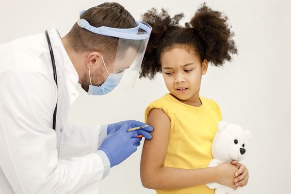 Male doctor in white medical gown is going to do vaccination. Man wearing white medical gown and face mask. Girl wearing yellow t-shirt and holding a bear toy in hands.