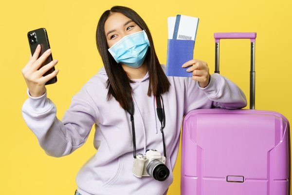 Happy korean woman taking selfie with passport and flight tickets, going on vacation, standing near big cute pink suitcase, yellow background.